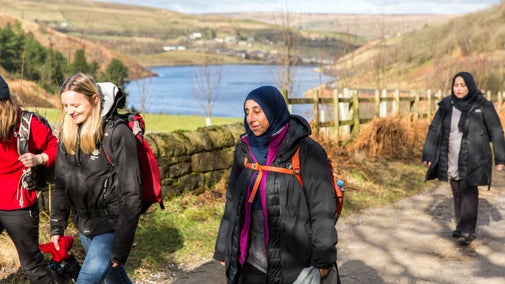 A group of four women along a path, in the background, down a hill is a reservoir